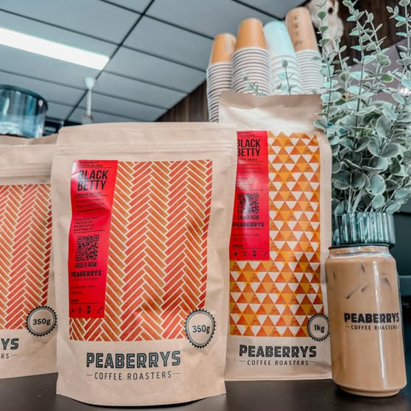 Three bags of Peaberrys Coffee Roasters in kraft paper packaging with orange and red geometric patterns are displayed on a counter next to a jar of iced coffee, with stacks of paper cups and a potted plant in the background.